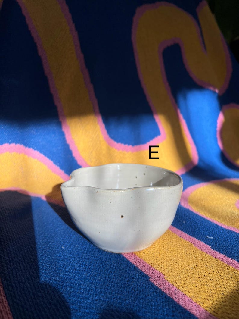 May include: A small, off-white ceramic bowl with a wavy rim. The bowl has a speckled appearance and a small spout. The background is a blue, yellow, and pink patterned fabric.
