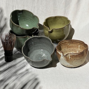 May include: A collection of four ceramic matcha bowls with scalloped rims and pouring spouts, featuring varied glaze colors including sage green, olive, slate gray, and speckled beige, accompanied by a dark brown bamboo tea whisk.