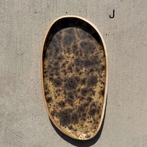 May include: An oval-shaped tray with an irregular, organic edge. The interior surface features a mottled, speckled pattern in shades of dark brown, tan, and cream, resembling a natural burl wood grain or stone texture. The letter J is visible.