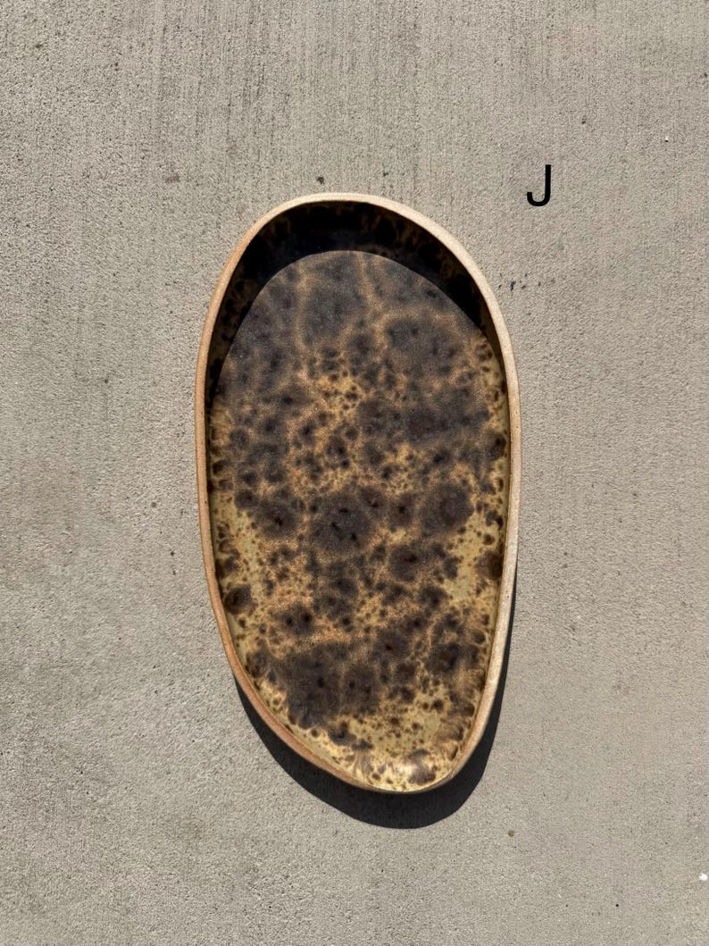 May include: An oval-shaped tray with an irregular, organic edge. The interior surface features a mottled, speckled pattern in shades of dark brown, tan, and cream, resembling a natural burl wood grain or stone texture. The letter J is visible.