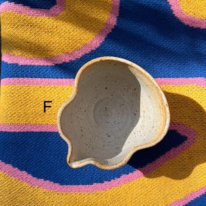 May include: A handmade, off-white ceramic bowl with a unique, irregular shape and a small spout. The bowl has a speckled texture and a light brown rim. The bowl is set against a colorful background of blue, yellow, and pink.