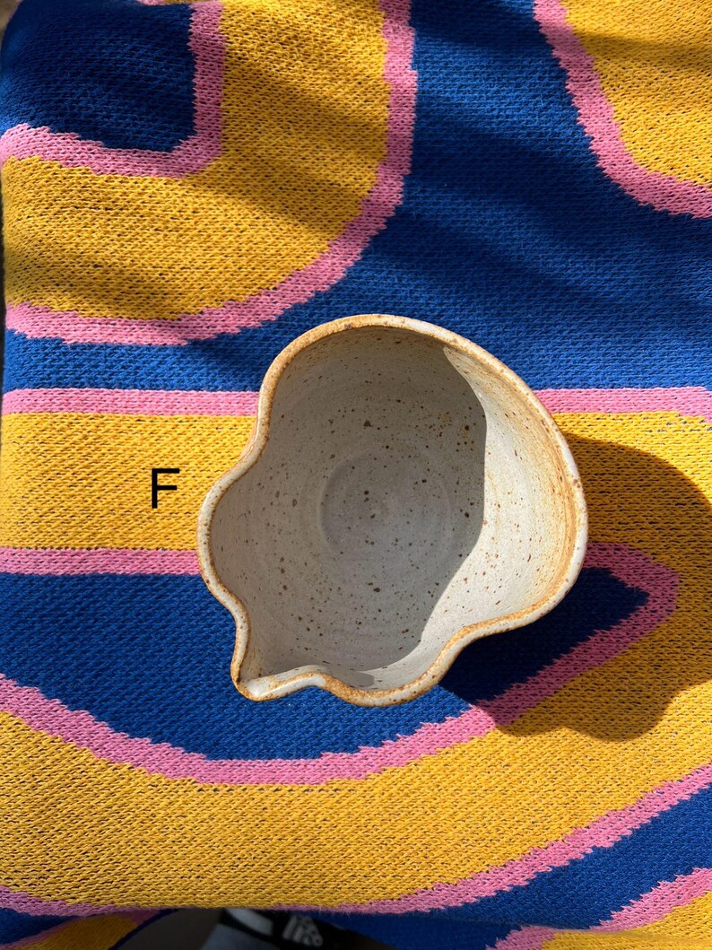 May include: A handmade, off-white ceramic bowl with a unique, irregular shape and a small spout. The bowl has a speckled texture and a light brown rim. The bowl is set against a colorful background of blue, yellow, and pink.
