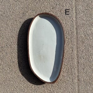 May include: An oval-shaped ceramic tray with a smooth, off-white glazed interior and a dark, textured brown rim. The tray features an irregular, organic edge and is marked with the letter E in the upper right corner of the composition.