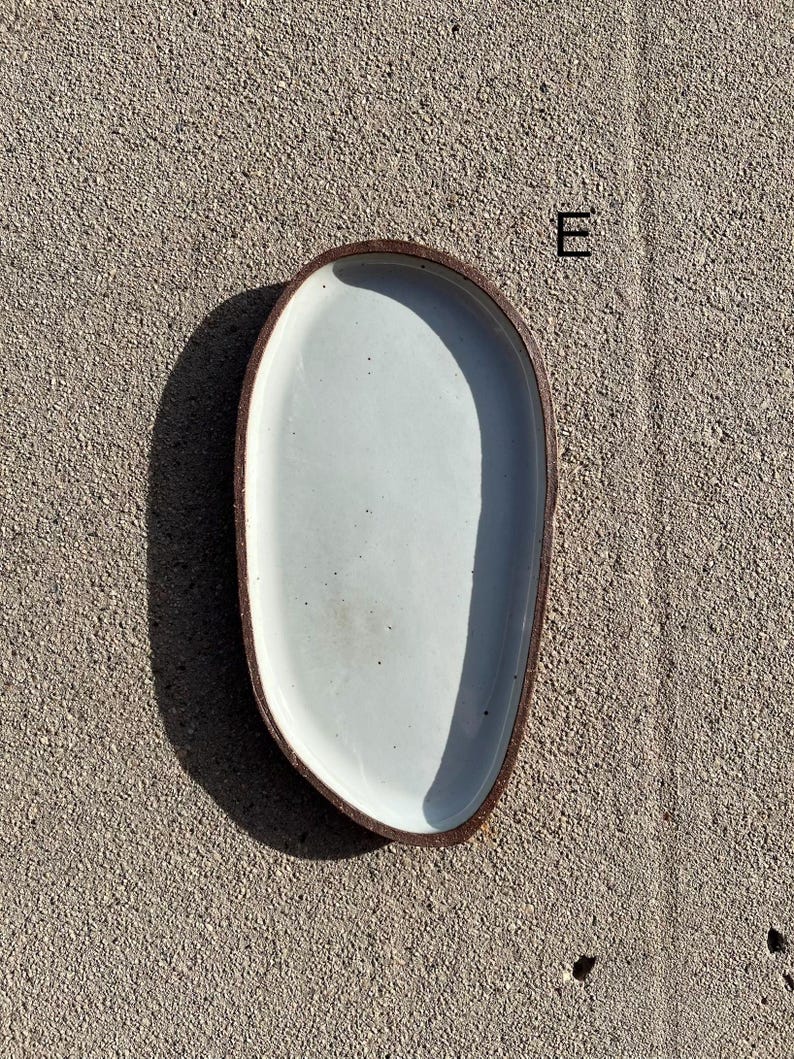 May include: An oval-shaped ceramic tray with a smooth, off-white glazed interior and a dark, textured brown rim. The tray features an irregular, organic edge and is marked with the letter E in the upper right corner of the composition.