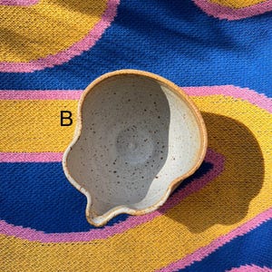 May include: A handmade ceramic bowl with a unique, irregular shape. The bowl has a speckled, off-white interior and a light brown rim. The letter "B" is visible on the side. The bowl is set against a colorful blue, yellow, and pink striped background.