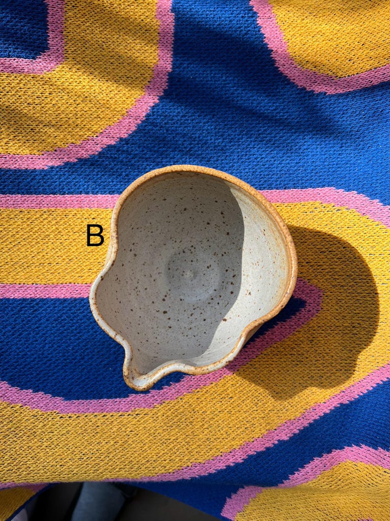 May include: A handmade ceramic bowl with a unique, irregular shape. The bowl has a speckled, off-white interior and a light brown rim. The letter "B" is visible on the side. The bowl is set against a colorful blue, yellow, and pink striped background.