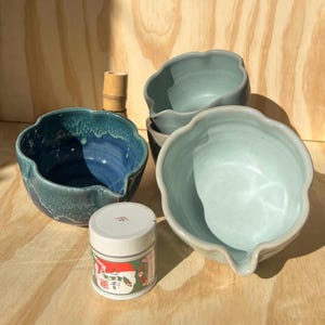 May include: A collection of ceramic matcha bowls in shades of blue and green, with a small container of matcha powder. The bowls have a unique, organic shape with a spout. A bamboo whisk is also visible.