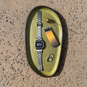 May include: An oval olive green ceramic tray with a dark rim containing a silver digital wristwatch with a metal link band, a small box of matches with dark tips, two silver rings, and a small brass-colored spherical object with a thin stem.