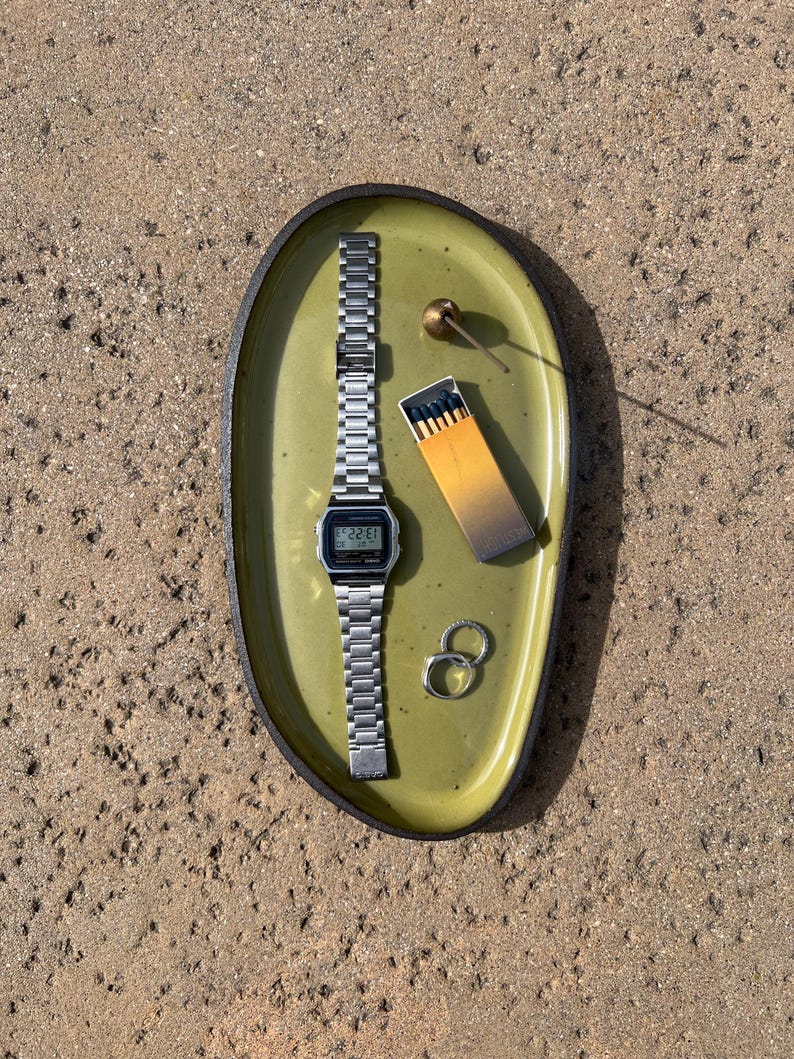 May include: An oval olive green ceramic tray with a dark rim containing a silver digital wristwatch with a metal link band, a small box of matches with dark tips, two silver rings, and a small brass-colored spherical object with a thin stem.