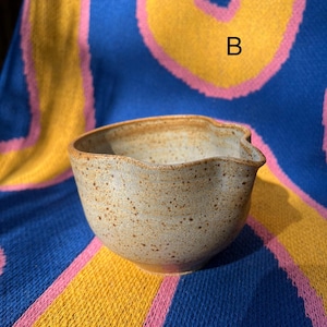 May include: A speckled, beige ceramic bowl with a unique, wavy rim and a pouring spout. The bowl is set against a colorful background of blue, yellow, and pink fabric. The letter "B" is visible on the fabric.