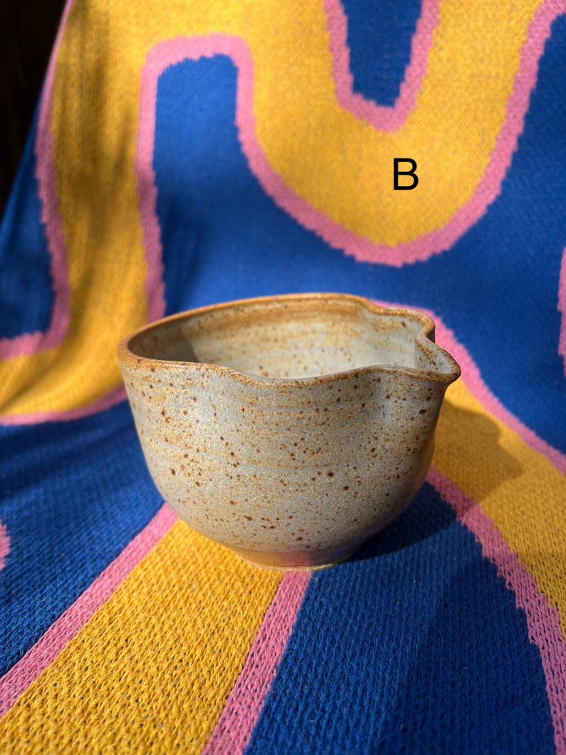 May include: A speckled, beige ceramic bowl with a unique, wavy rim and a pouring spout. The bowl is set against a colorful background of blue, yellow, and pink fabric. The letter "B" is visible on the fabric.