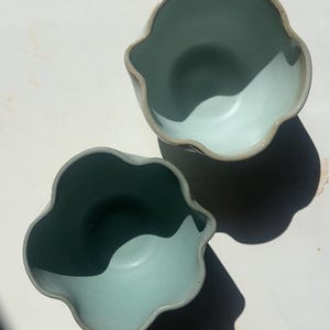 May include: Two light blue ceramic bowls with a wavy rim and a contrasting light brown edge. The bowls are placed on a white surface, casting a shadow. The bowls have a smooth, glossy finish.