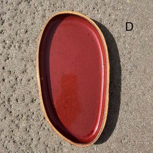 May include: An oval-shaped ceramic tray with a glossy deep red interior glaze and a textured, unglazed tan rim. The tray features an irregular, organic edge and is marked with the letter D in the upper right corner, showcasing a speckled finish.
