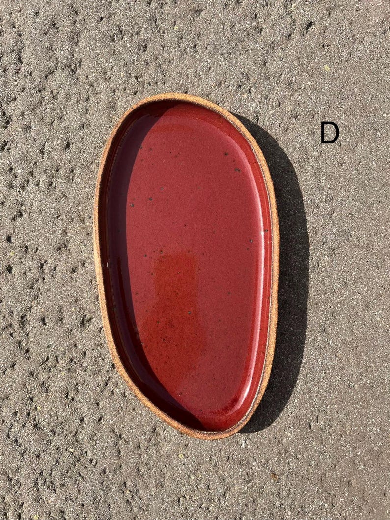 May include: An oval-shaped ceramic tray with a glossy deep red interior glaze and a textured, unglazed tan rim. The tray features an irregular, organic edge and is marked with the letter D in the upper right corner, showcasing a speckled finish.