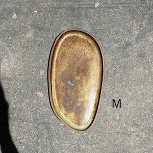 May include: An oval-shaped ceramic tray with a speckled brown and tan glaze and a dark brown rim. The tray features an irregular, organic edge and is marked with the letter M in black text on the right side of the object's surface.