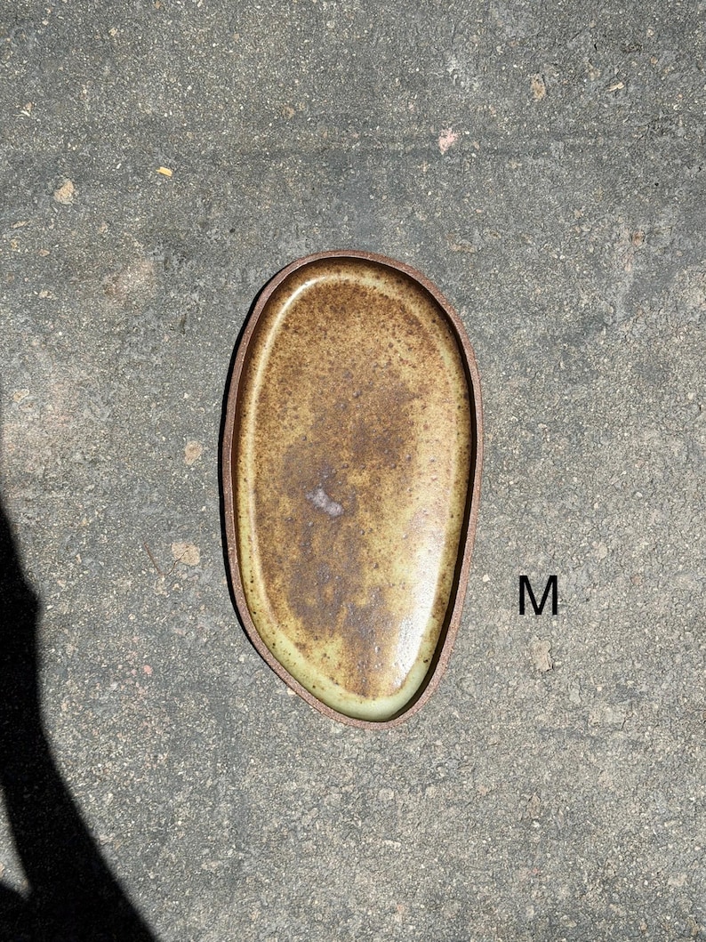 May include: An oval-shaped ceramic tray with a speckled brown and tan glaze and a dark brown rim. The tray features an irregular, organic edge and is marked with the letter M in black text on the right side of the object's surface.