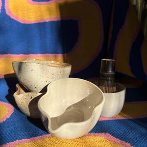 May include: A collection of handmade ceramic bowls in varying sizes and shapes. The bowls are off-white with speckled details and a natural, organic aesthetic. A bamboo whisk sits in a white ceramic bowl. The background is a colorful textile.