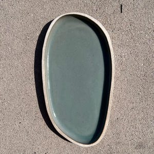 May include: An oval-shaped ceramic tray with an irregular, organic edge. The interior surface features a smooth, matte sage green glaze, while the outer rim and base remain in a contrasting, unglazed light beige clay finish. The number 1 is visible.
