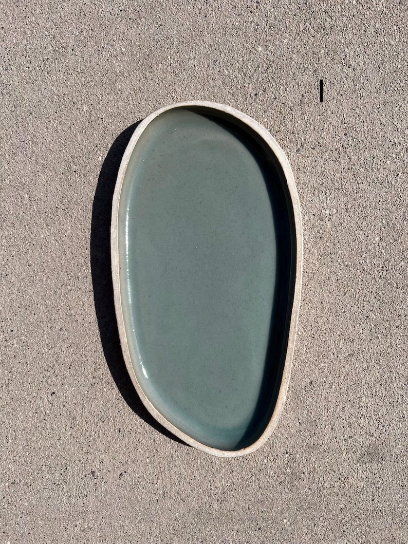 May include: An oval-shaped ceramic tray with an irregular, organic edge. The interior surface features a smooth, matte sage green glaze, while the outer rim and base remain in a contrasting, unglazed light beige clay finish. The number 1 is visible.