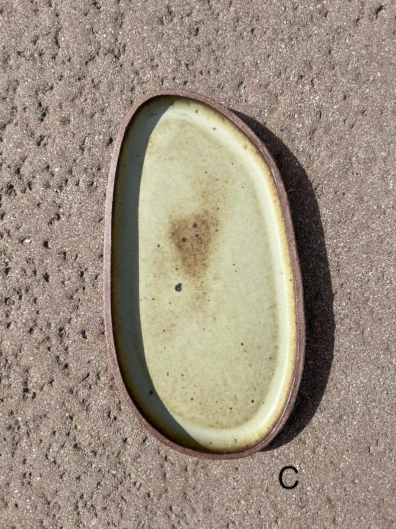 May include: An oval ceramic tray with a light sage green interior glaze featuring dark brown speckles and a central mottled brown patch. The tray has a raised, dark brown, textured rim that contrasts with the smooth, lighter-colored center.