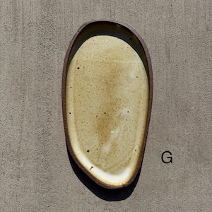 May include: An oval-shaped ceramic tray with an irregular, organic edge. The interior surface features a speckled, light beige glaze with subtle white highlights, while the outer rim is a dark, matte brown. The letter G is printed to the right.