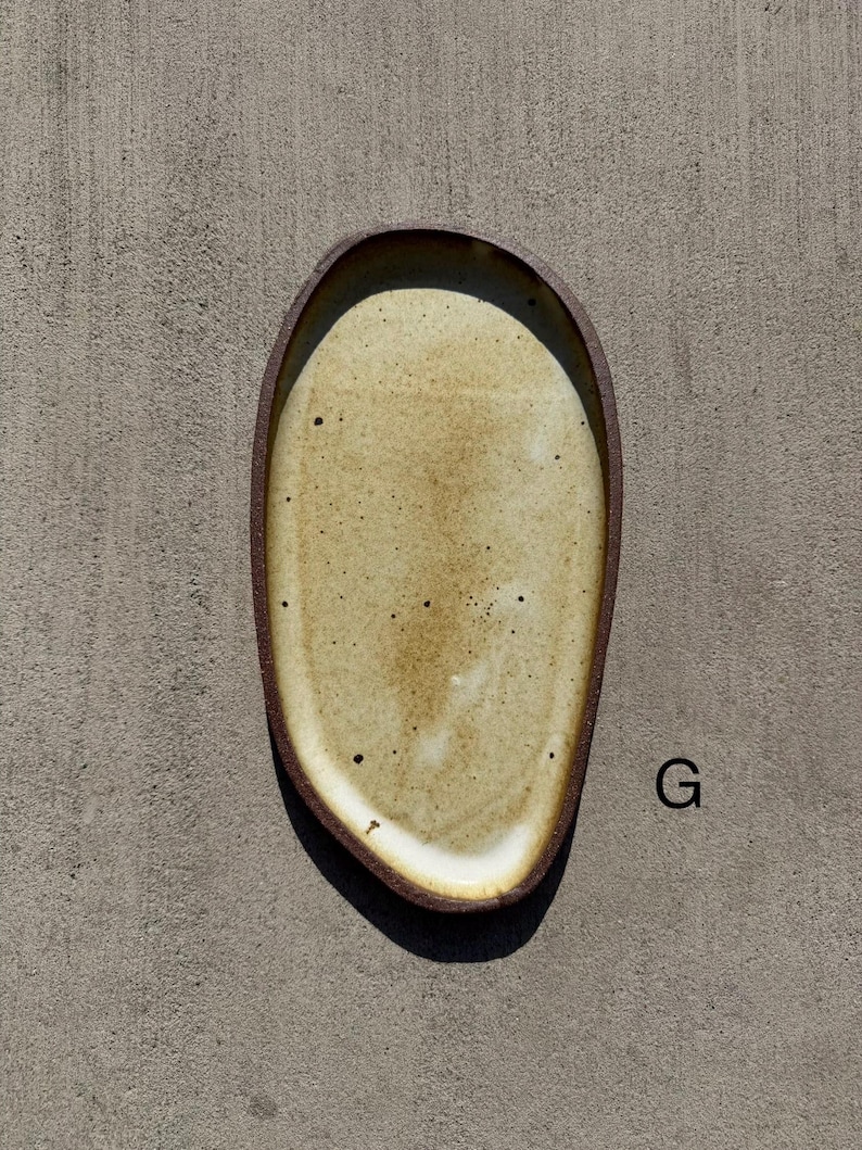 May include: An oval-shaped ceramic tray with an irregular, organic edge. The interior surface features a speckled, light beige glaze with subtle white highlights, while the outer rim is a dark, matte brown. The letter G is printed to the right.