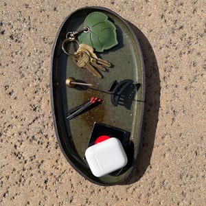 May include: An oval-shaped dark green ceramic tray containing a set of metal keys with a green leaf-shaped keychain, a small gold-toned sphere on a thin stick, a black hair claw clip, three matches with red tips, and a white wireless earbud case.