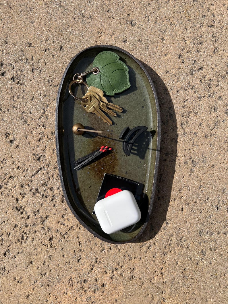 May include: An oval-shaped dark green ceramic tray containing a set of metal keys with a green leaf-shaped keychain, a small gold-toned sphere on a thin stick, a black hair claw clip, three matches with red tips, and a white wireless earbud case.