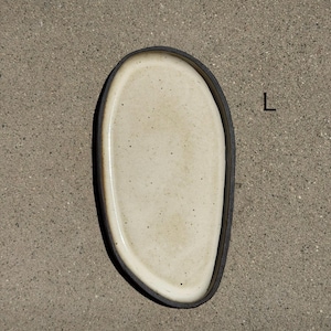 May include: An oval ceramic tray with a speckled off-white interior and a dark brown, textured outer rim. The tray features an irregular, organic shape and is marked with the letter L in black text on the right side of the object.