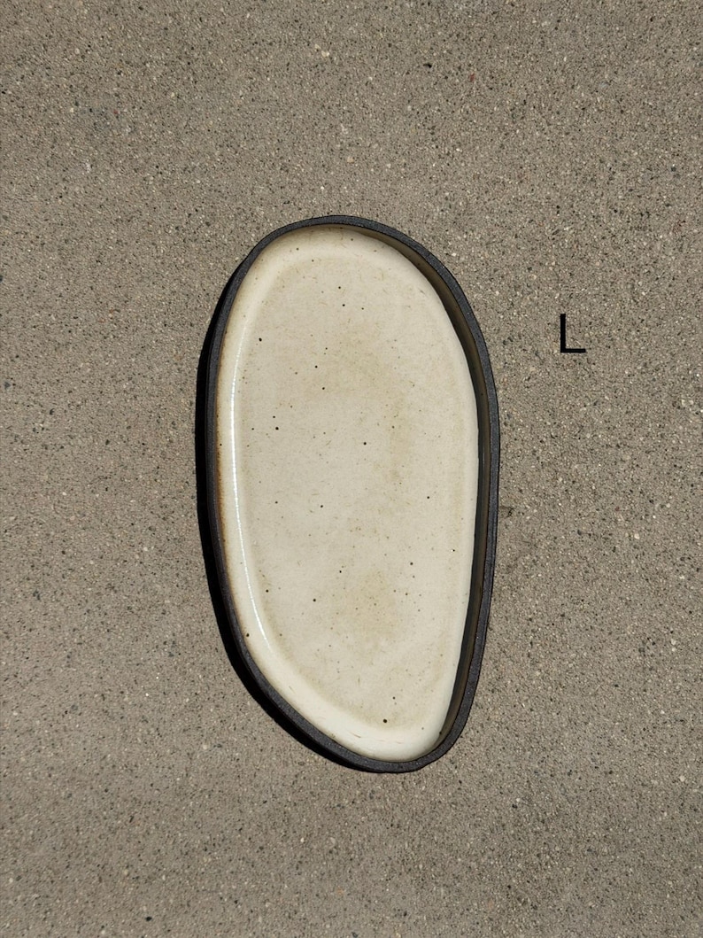 May include: An oval ceramic tray with a speckled off-white interior and a dark brown, textured outer rim. The tray features an irregular, organic shape and is marked with the letter L in black text on the right side of the object.