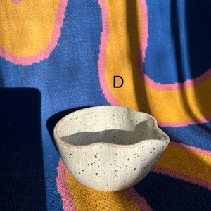 May include: A small, off-white ceramic bowl with a spout and speckled brown details. The bowl is set against a colorful background of blue, yellow, and pink. The letter "D" is visible in the background.