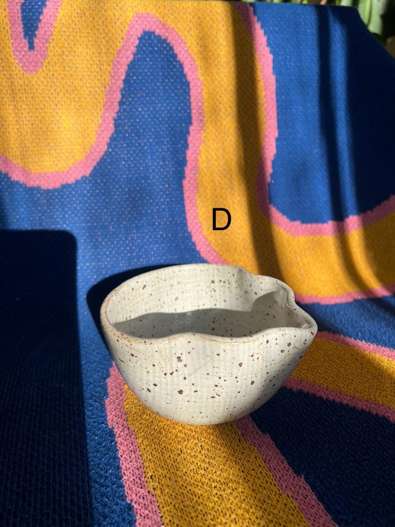 May include: A small, off-white ceramic bowl with a spout and speckled brown details. The bowl is set against a colorful background of blue, yellow, and pink. The letter "D" is visible in the background.