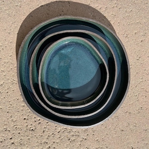 May include: A set of three nested ceramic plates with irregular, organic shapes. The plates feature a deep, glossy teal and dark blue glaze with a smooth finish, contrasted by a dark, matte, textured rim that defines their unique, non-circular edges.