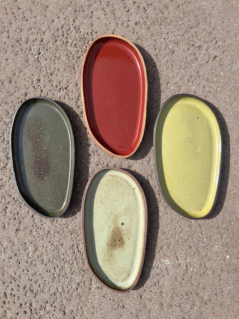 May include: Four oval-shaped ceramic plates with irregular, organic edges. The set includes one deep red plate, one dark forest green plate, one light lime green plate, and one pale cream-colored plate with a speckled finish and dark rims.