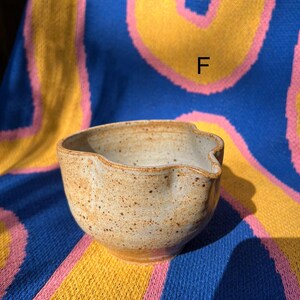May include: A handmade, speckled ceramic bowl with a pouring spout. The bowl is a light tan color with brown speckles and a smooth, rounded shape. The bowl is sitting on a blue, yellow, and pink patterned surface.
