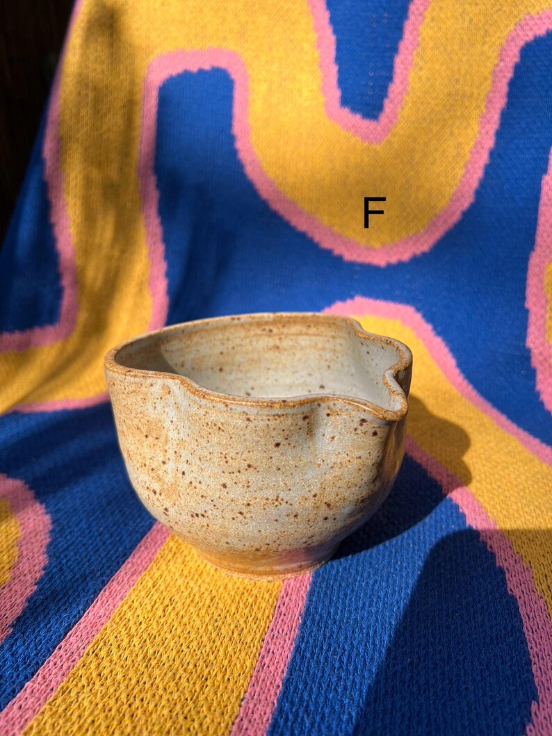 May include: A handmade, speckled ceramic bowl with a pouring spout. The bowl is a light tan color with brown speckles and a smooth, rounded shape. The bowl is sitting on a blue, yellow, and pink patterned surface.