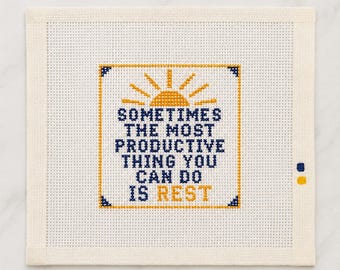 Könnte beinhalten: Ein quadratisches Stickleinen mit einem Kreuzstichmotiv, das oben ein gelbes Sonnensymbol und den dunkelblauen Text "SOMETIMES THE MOST PRODUCTIVE THING YOU CAN DO IS REST" zeigt, wobei das Wort "REST" gelb ist, umrahmt von einem dünnen gelben Rand.