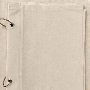 Puede incluir: Una pila de hojas de tela rectangulares de color blanco roto hechas de un material de tejido liso. Las hojas están unidas en el lado izquierdo por dos anillas de metal plateado que pasan a través de ojales reforzados en los bordes.