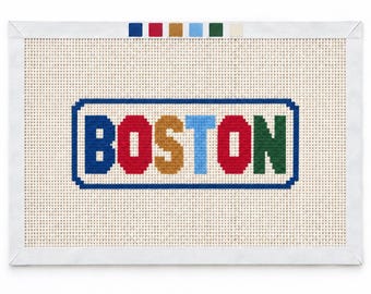 Könnte beinhalten: Eine rechteckige Stickleinwand mit dem Wort BOSTON, das in einer fetten, mehrfarbigen Schrift innerhalb eines blauen rechteckigen Rahmens gestickt ist. Die Buchstaben sind blau, rot, gold, hellblau, rot und grün. Ein weißer Stoffrand umgibt das Gitter.