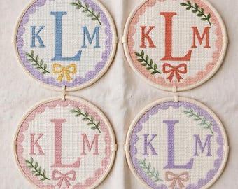 Könnte beinhalten: Vier kreisförmige Kreuzstich-Stickrahmen mit dem Monogramm KLM. Jedes Stück hat einen gewellten Rand in Pastellfarben, einen großen zentralen Buchstaben L, kleinere Buchstaben K und M, eine dekorative Schleife und grüne Blattzweige auf weißem Stoff.