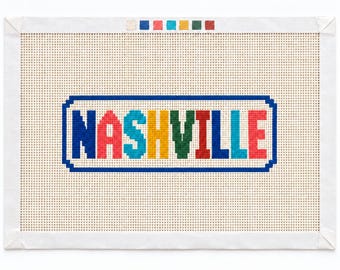 Könnte beinhalten: Eine rechteckige Stickleinwand mit dem Wort NASHVILLE in mehrfarbigen Blockbuchstaben, wobei jeder Buchstabe einen anderen Farbton aufweist. Der Text ist von einem blauen rechteckigen Rahmen umgeben und liegt auf einem Gitterhintergrund.