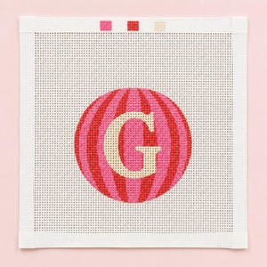 May include: A square needlepoint canvas featuring a central circular design with alternating vertical stripes in shades of pink and red. A large, cream-colored capital letter G is centered within the circle. Three small color swatches are at the top.