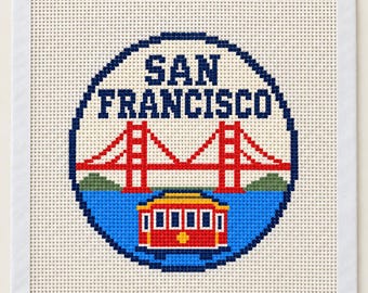 Könnte beinhalten: Ein quadratisches Stickbild mit einem kreisförmigen Motiv und dem Text SAN FRANCISCO in Dunkelblau. Das Design zeigt eine rote Golden Gate Bridge, grüne Hügel und eine rote Straßenbahn auf blauem Wasser. Oben befinden sich sechs kleine Farbquadrate.