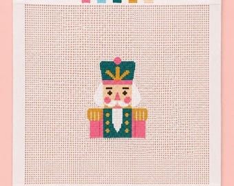 Könnte beinhalten: Ein quadratisches Stickbild mit einem zentrierten Nussknacker-Motiv in Petrol, Pink, Gold und Weiß. Der Nussknacker trägt einen hohen petrolfarbenen Hut mit goldenem Emblem und eine passende Uniform. Fünf kleine farbige Quadrate sind oben ausgerichtet.