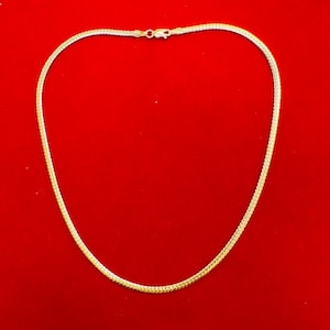May include: A gold-coloured chain necklace with a woven design. The necklace is displayed against a solid red background. The clasp is visible at the top of the necklace.
