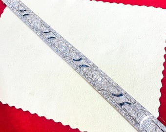 May include: A silver-toned bracelet with a repeating pattern of clear and dark blue stones. The bracelet is rectangular and lies diagonally across a white surface, set against a red background.