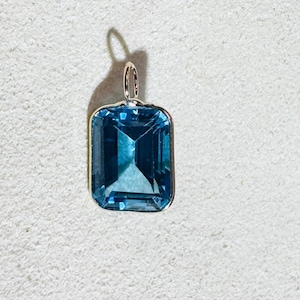 May include: A rectangular, blue gemstone pendant with a silver-colored bail. The gemstone has a faceted cut, reflecting light. The pendant is set in a silver-colored metal frame. The background is a textured, off-white surface.
