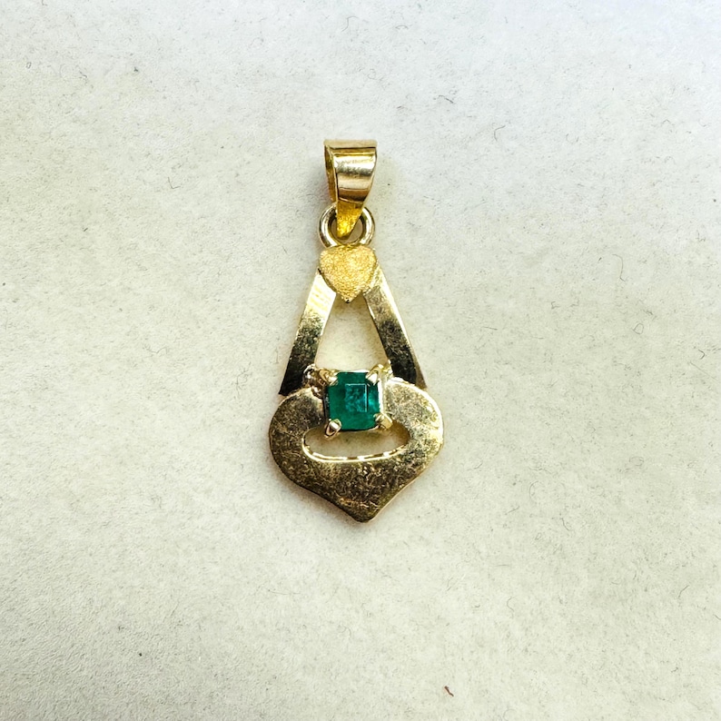 May include: A gold pendant with a heart-shaped design and a square emerald gemstone. The pendant has a small loop at the top for a chain. The emerald is a vibrant green color, and the gold has a slightly aged appearance.