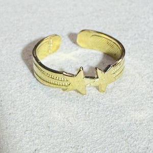 Puede incluir: Un anillo abierto de tono dorado que presenta dos estrellas prominentes en el centro. La banda tiene un diseño texturizado y acanalado que se extiende desde las estrellas hacia la abertura posterior, creando un patrón continuo alrededor.