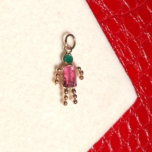Peut inclure: Un pendentif doré en forme de personne. La tête est une pierre précieuse verte, le corps est une pierre précieuse rose et les bras et les jambes sont de petites perles dorées. Le pendentif est sur une surface blanche avec un fond rouge.